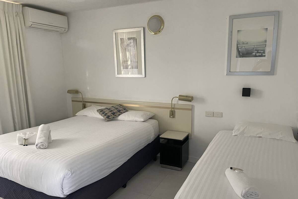 A hotel room with two beds, a bedside table, an air conditioning unit, and two framed pictures on the wall.