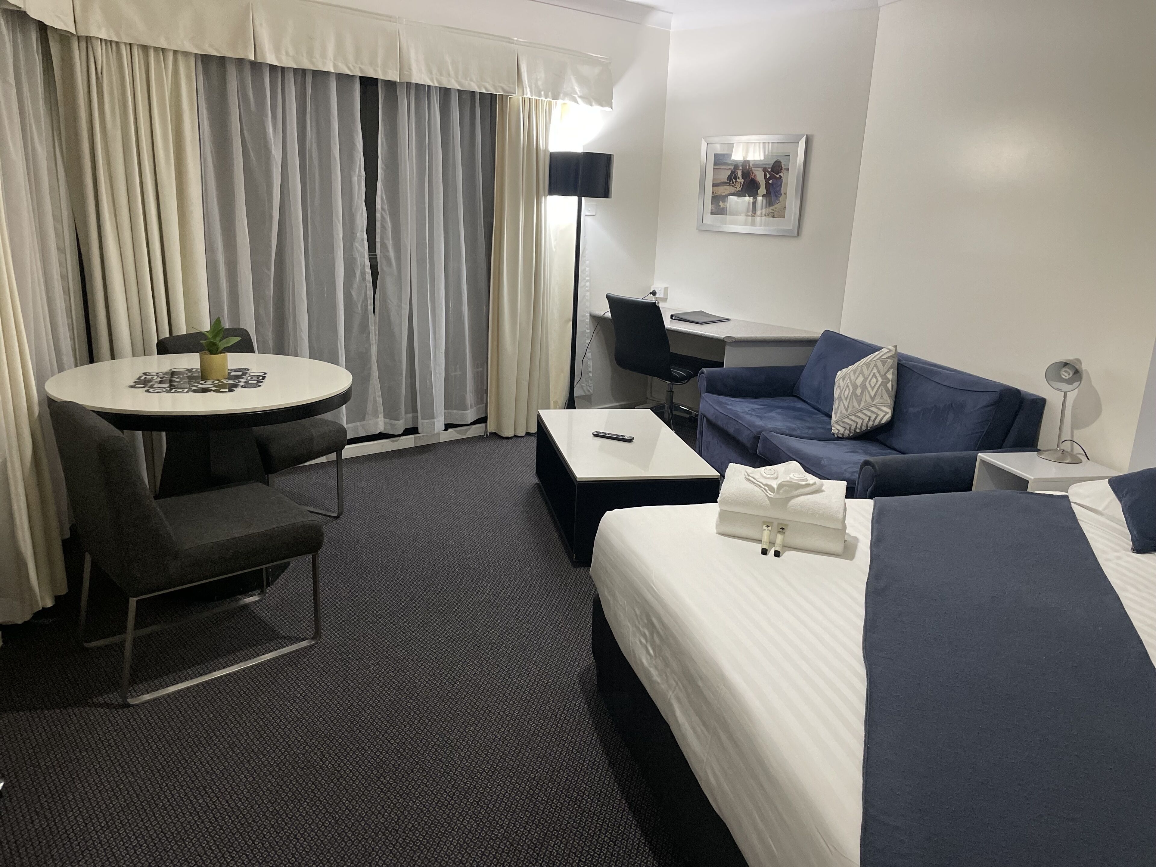 A hotel room with a bed, a sofa, a desk, a chair, a small table with a plant, and a window with curtains.
