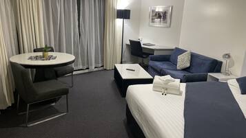 A hotel room with a bed, a sofa, a desk, a chair, a small table with a plant, and a window with curtains.