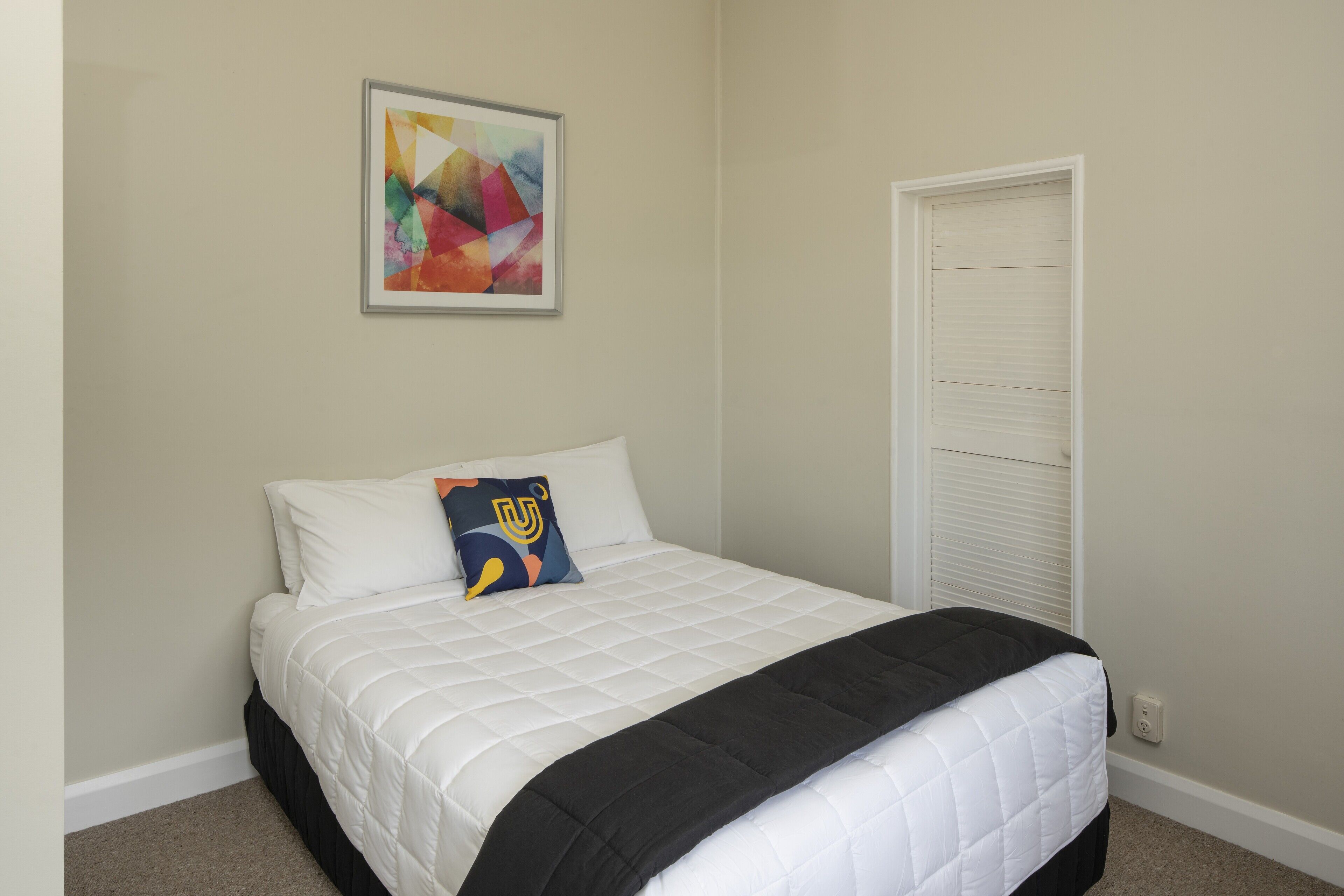 A neatly made bed with a white quilt and a black throw blanket, a colorful abstract painting on the wall, and a closed white door.