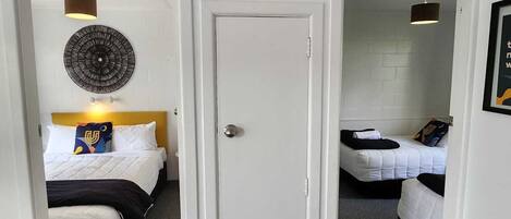 A hotel room with a bed, a bedside table, a wall-mounted lamp, and a door leading to another room.