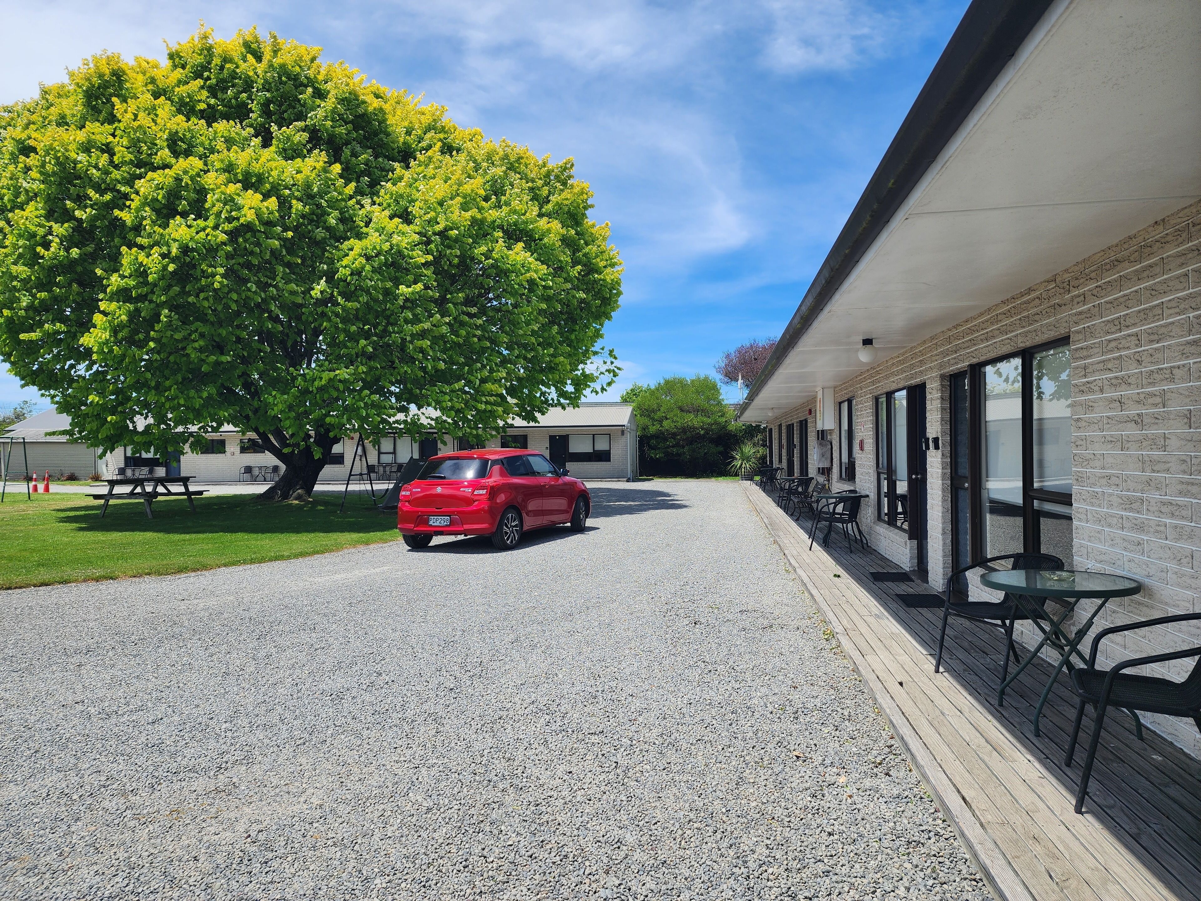 A red car parked in front of a building with a gravel driveway and a large tree.