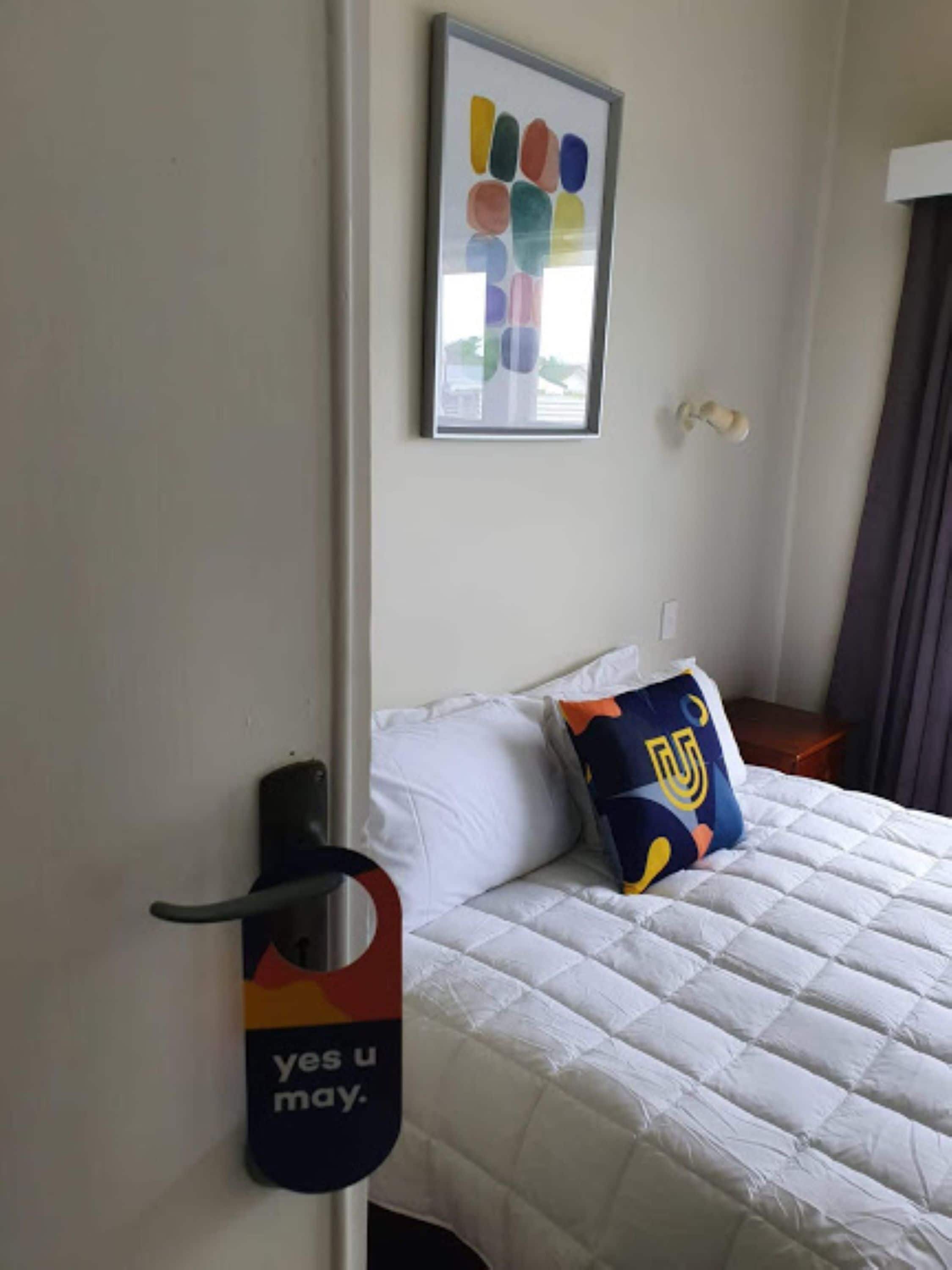 Ein Hotelzimmer mit einem Bett, einem Logo-Kissen, einem Wandbild und einem Türgriff.