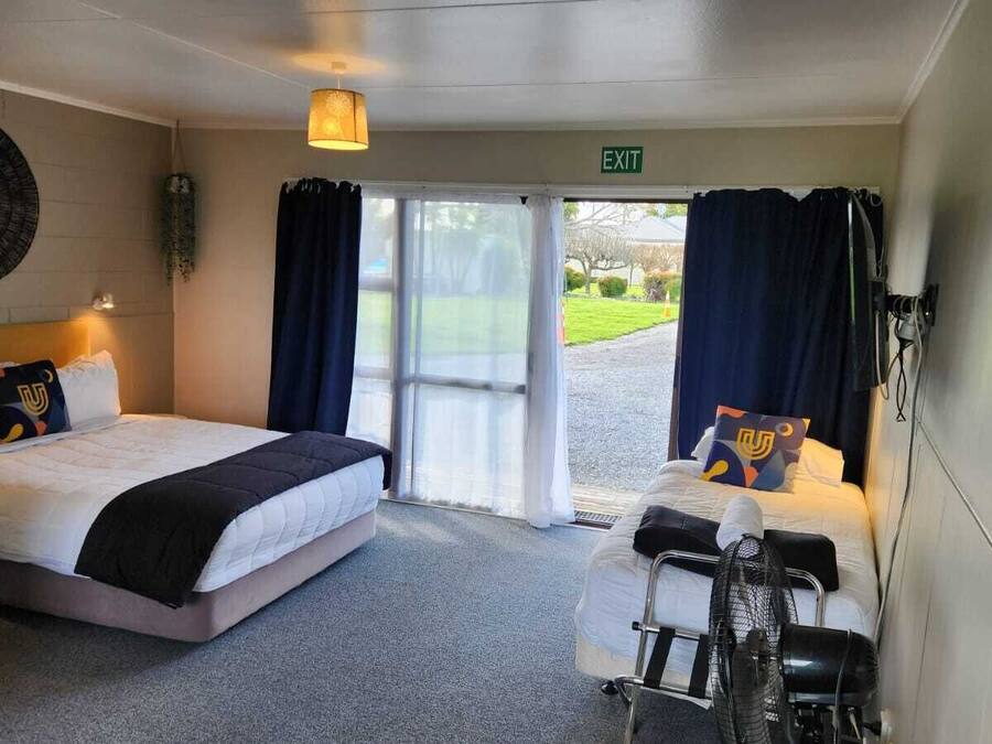 A hotel room with a bed, a desk, and a chair. There is a window with curtains, a wall clock, and an exit sign.