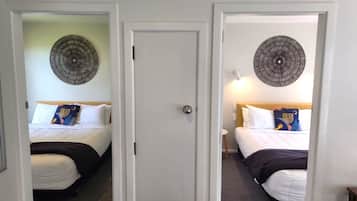Dois quartos de hotel lado a lado, separados por uma porta branca, cada um com uma cama, almofadas e um ornamento circular na parede.