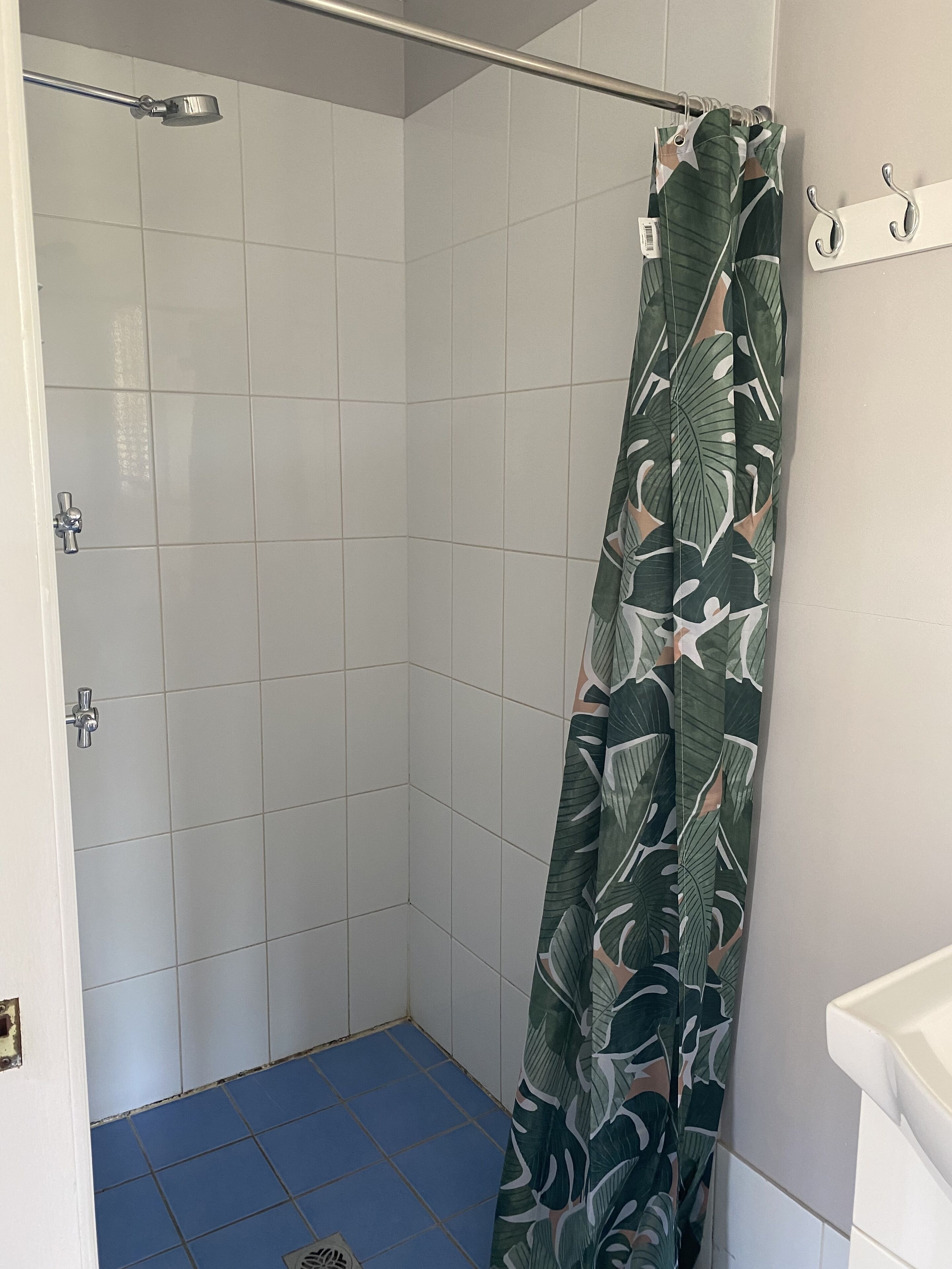 Ducha con azulejos blancos, piso azul y cortina de baño verde con estampado de hojas.