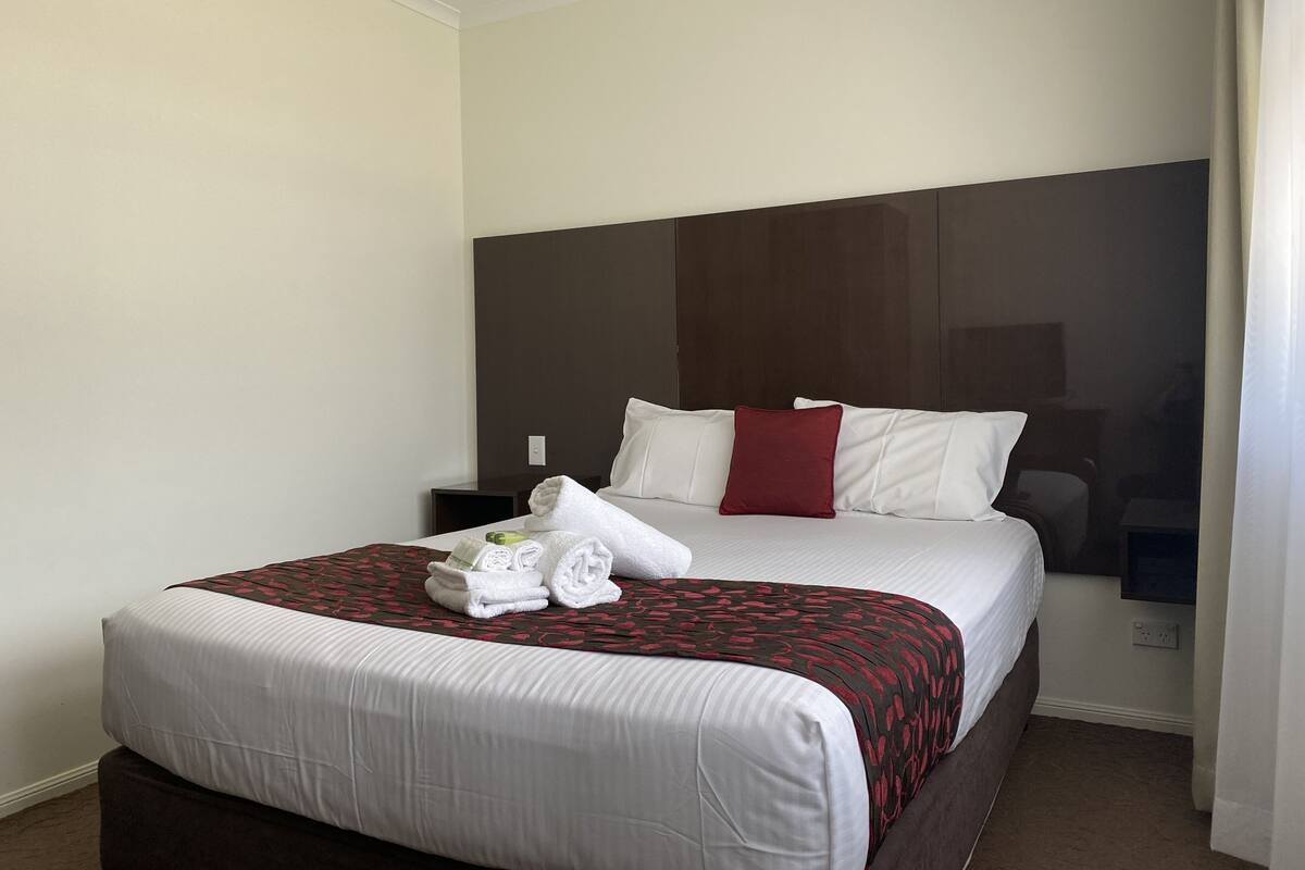 A hotel room with a large bed, a dark headboard, and a red accent pillow. There are two white bedside tables and a nightstand.