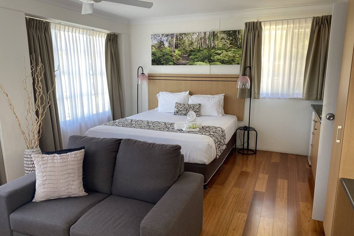A modern hotel room with a large bed, a grey sofa, a ceiling fan, and a painting of a forest on the wall.