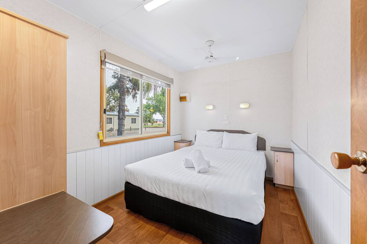 Une chambre d’hôtel avec un lit, des tables de chevet, un banc en bois, une fenêtre et un ventilateur de plafond.