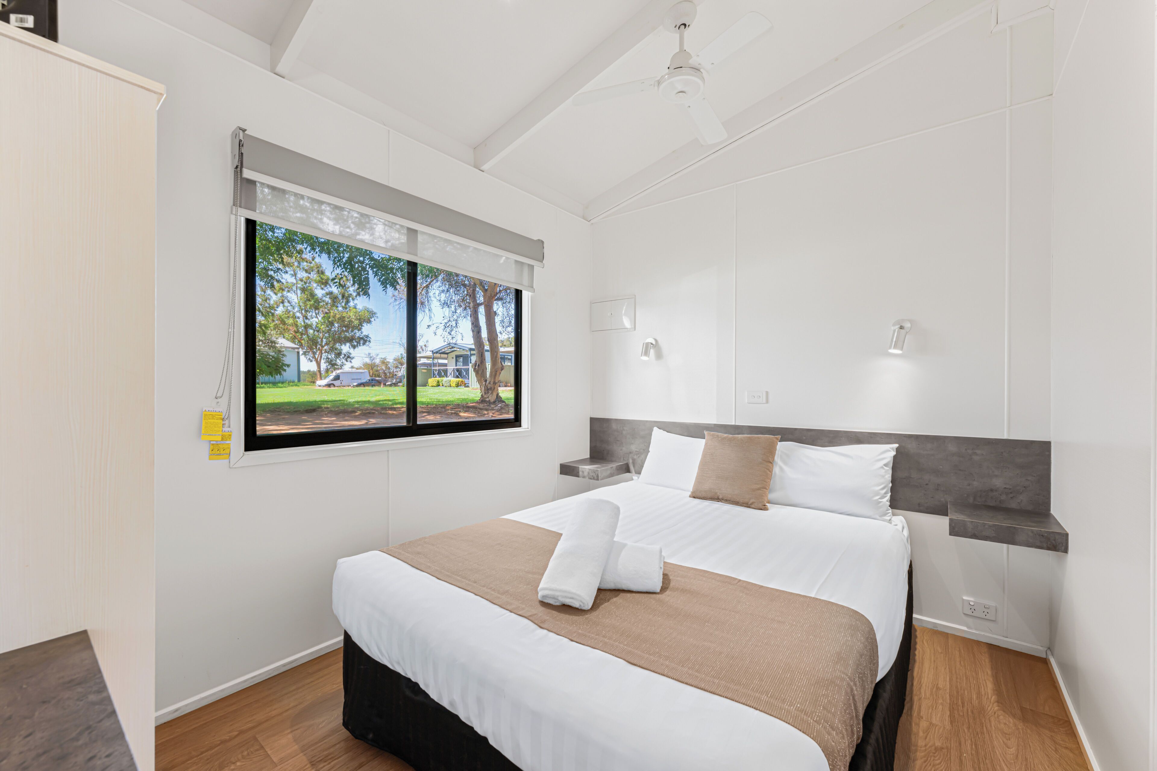 A hotel room with a large bed, a window with blinds, a ceiling fan, and a view of a residential area.