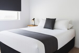 Una cama bien tendida con cubrecama blanco y gris oscuro, dos almohadas, una mesita de noche con una lámpara y una ventana al fondo.