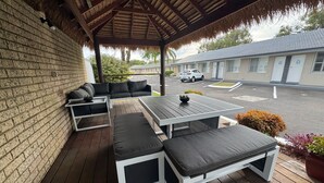 Uma área de estar coberta ao ar livre, com uma mesa de madeira e bancos, uma parede de tijolos e vista para uma rua com carros estacionados e edifícios.