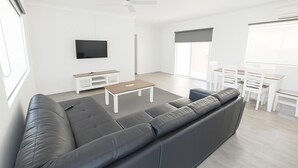 Uma sala de estar moderna com um sofá cinzento grande, uma mesa de centro, uma TV e uma área de jantar com móveis brancos.