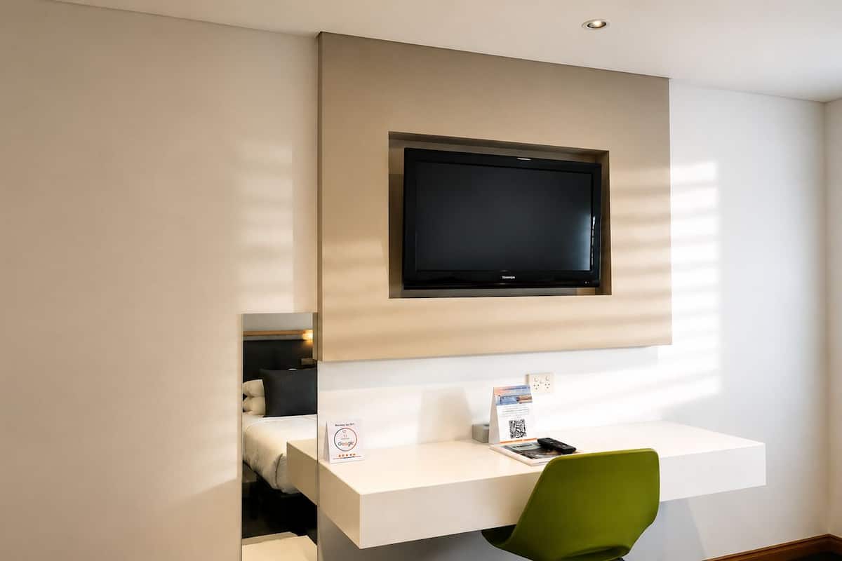 Ein Hotelzimmer mit montiertem Fernseher, einem Schreibtisch mit Stuhl und einem Bett.