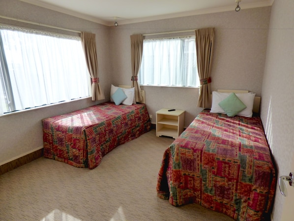 A room with two single beds, each with a patterned bedspread, a small bedside table, and a window with curtains. - Deco City Motor Lodge (Napier)