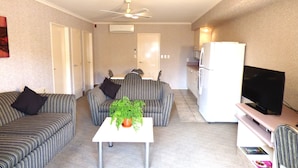 Un espacio habitable pequeño con sofás a rayas, una mesa central con una planta en maceta, un ventilador de techo, un refrigerador blanco y un televisor sobre un mueble.