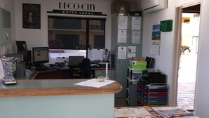 Un mostrador de recepción con una computadora, un calendario y un tablón de anuncios. Se ve una ventana con vista a una calle y coches estacionados.