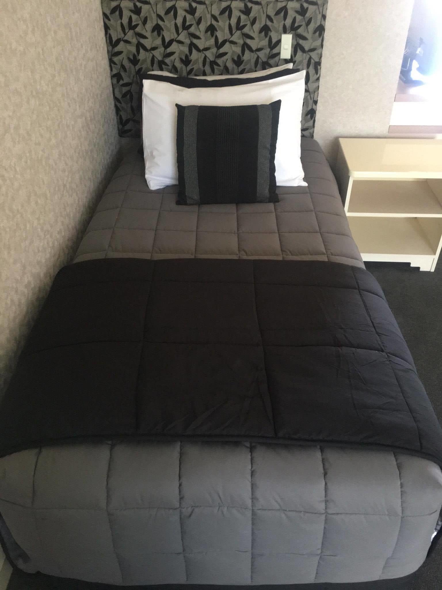 A bed with a grey quilt and pillows, a white bedside table, and a patterned wall.