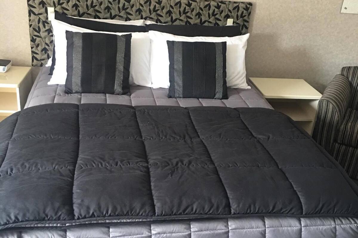 A neatly made bed with a dark quilt and pillows, a headboard with a leaf pattern, and a small bedside table with a lamp.
