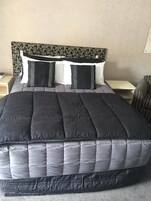 Una cama bien hecha con colcha oscura y almohadas, un cabecero con estampado de hojas y una mesita de noche con una lámpara.