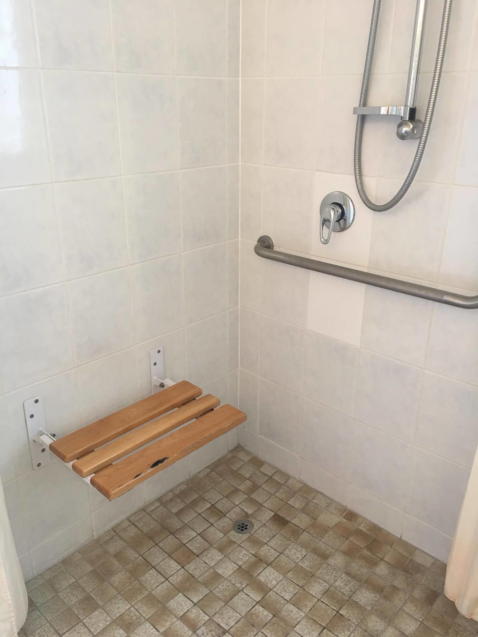 Zona de ducha con barra de apoyo, banco de madera y suelo de baldosas.