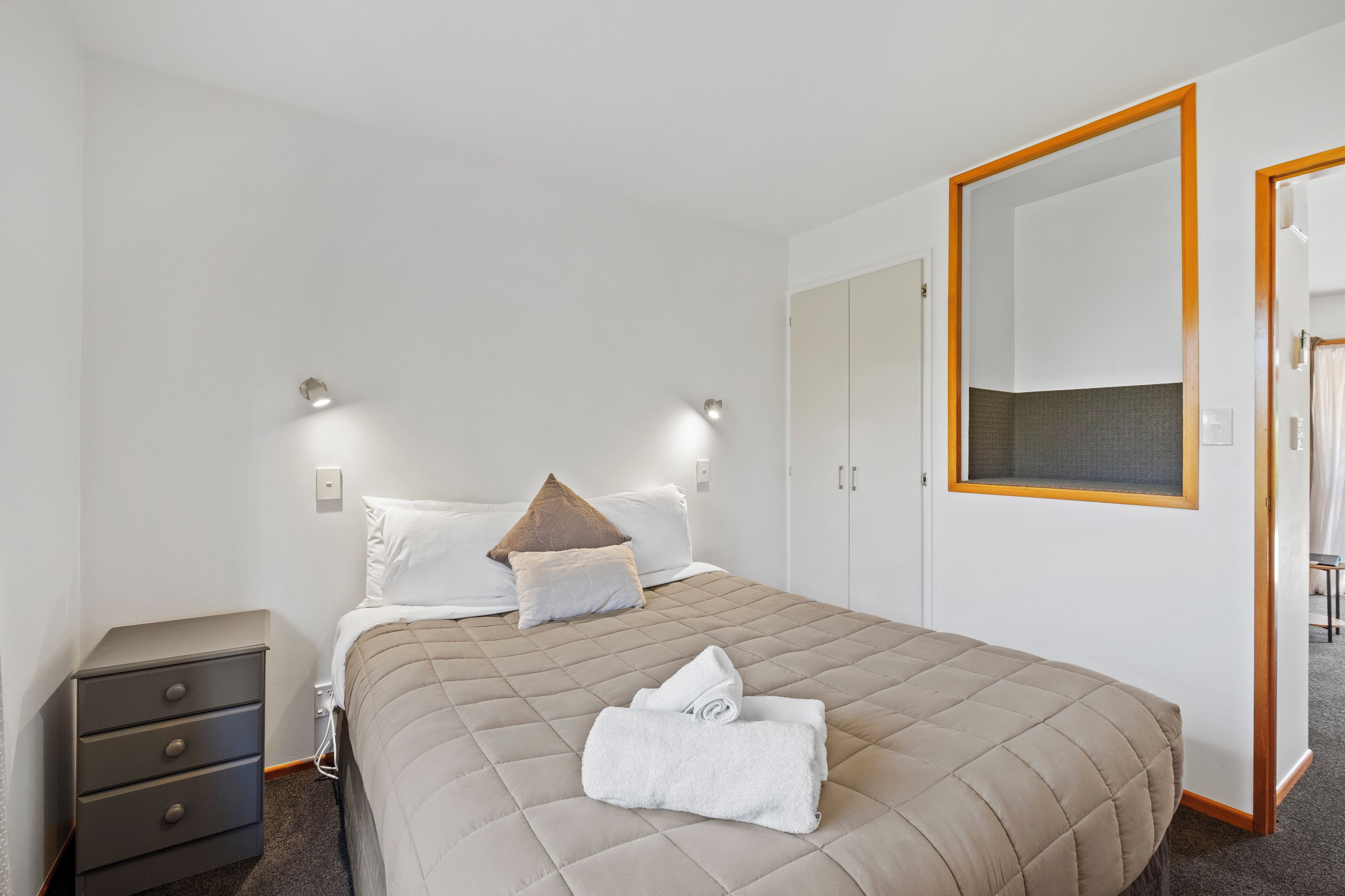 Un lit bien fait, recouvert d’une courtepointe, une table de chevet avec des tiroirs, un miroir fixé au mur et une armoire avec un miroir.