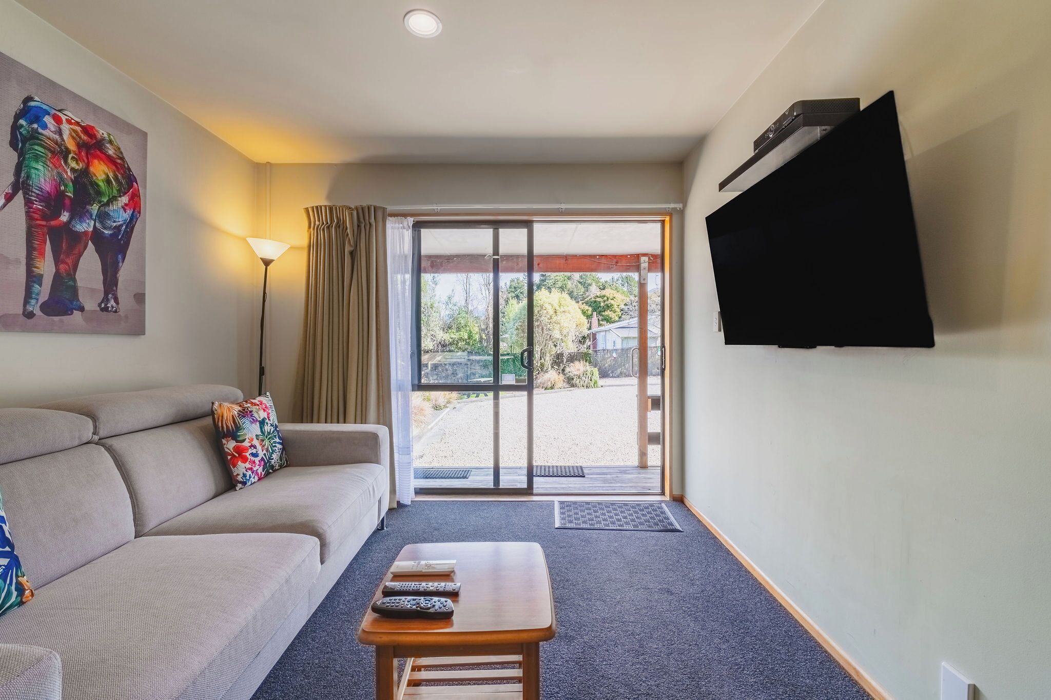 A living room with a sofa, a coffee table, a TV mounted on the wall, and a sliding glass door leading to an outdoor area.