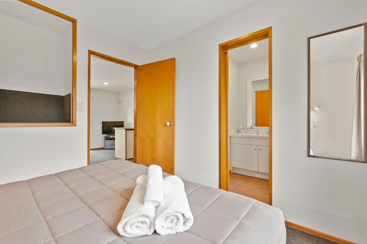 A hotel room with a bed, a bathroom mirror, and a door leading to a kitchenette.