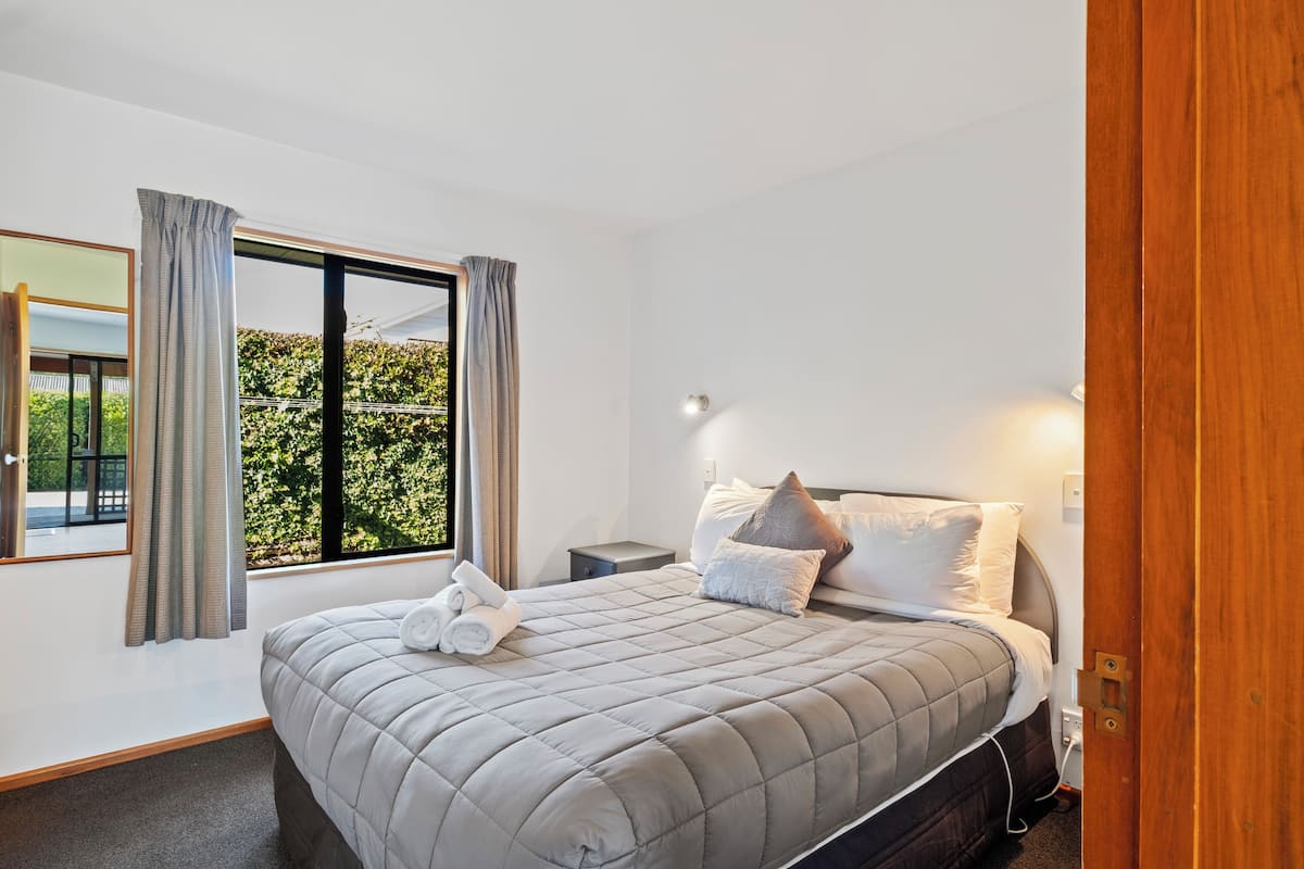 A modern bedroom with a large bed, a nightstand, a bedside lamp, and a view of greenery outside.