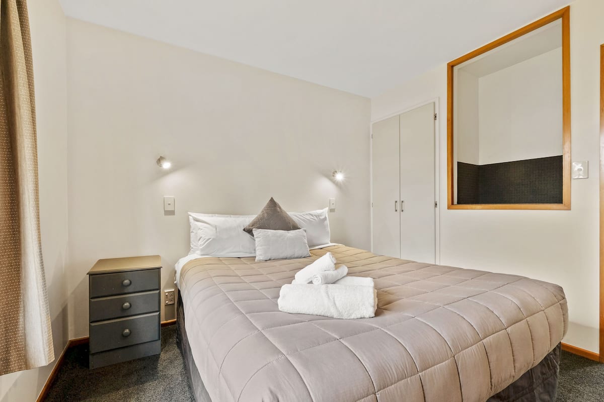 A neatly made bed with a quilted headboard, a bedside table with drawers, and a large mirror on the wall.