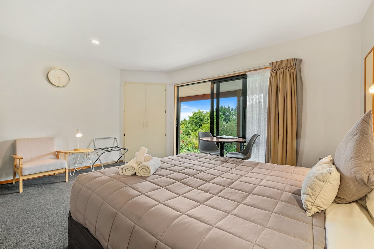 A hotel room with a large bed, a seating area with a table and chairs, a view of greenery through the window, and a door leading to an outdoor area.