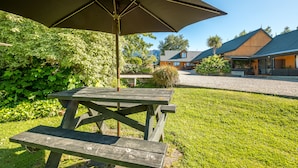 A picnic table with a large umbrella in a grassy area with a house in the background. - Alpine Garden Motel (Hanmer Springs)