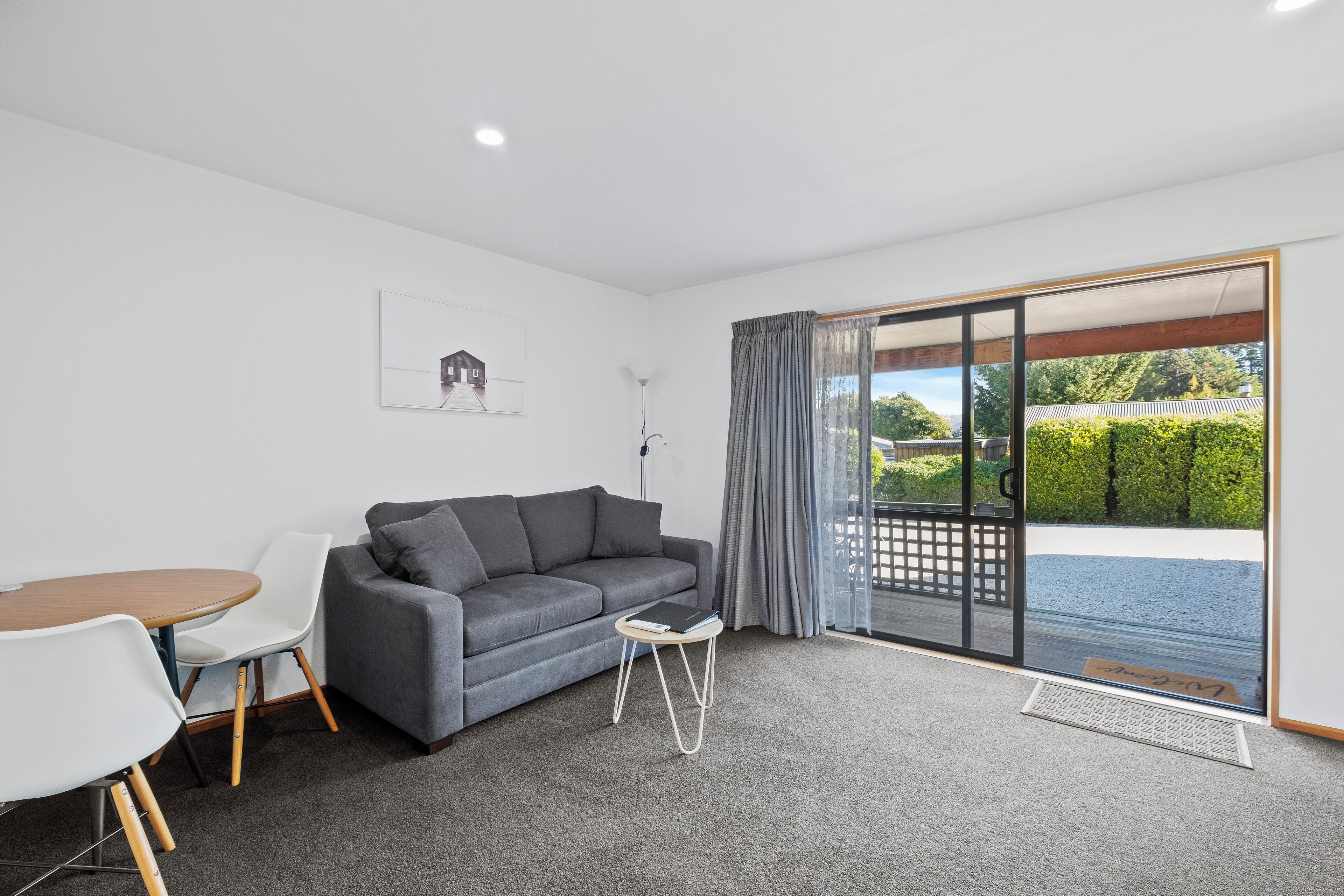A modern living room with a grey sofa, white chairs, a small round table, and a large sliding glass door leading to an outdoor area.