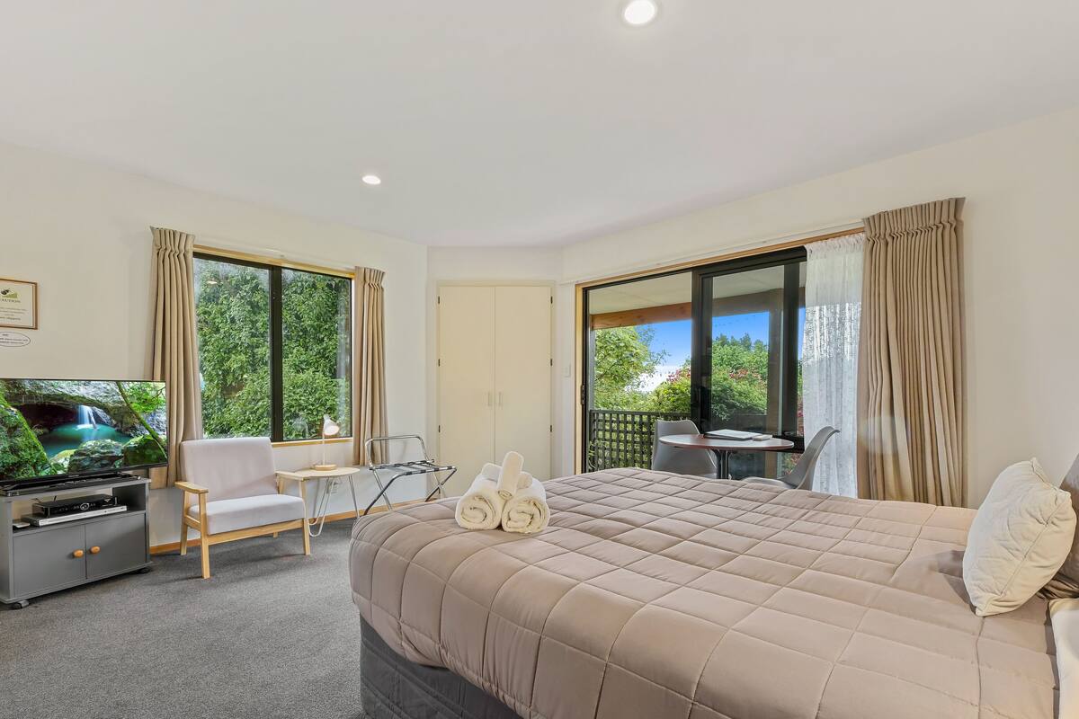 A hotel room with a large bed, a TV, a desk, and a balcony with a view of greenery.
