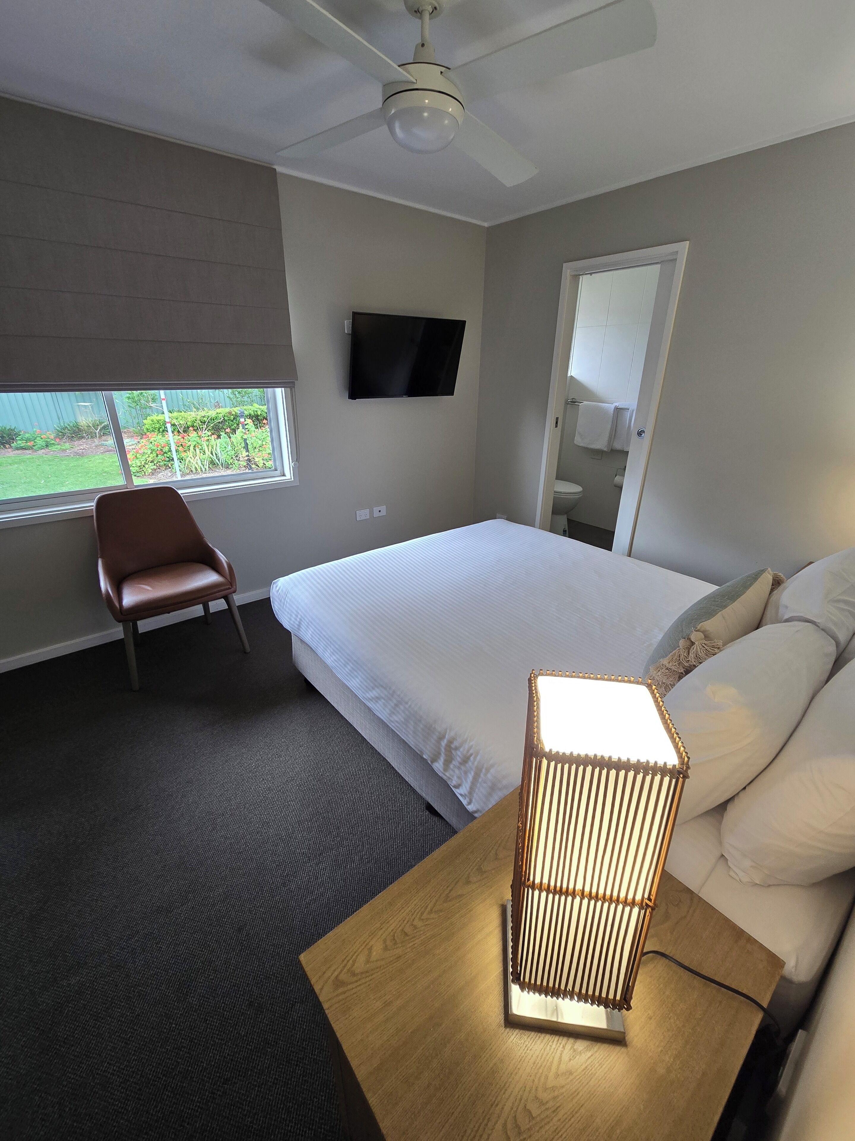 A hotel room with a bed, a chair, a TV, a ceiling fan, a window with a view, and a bedside lamp.