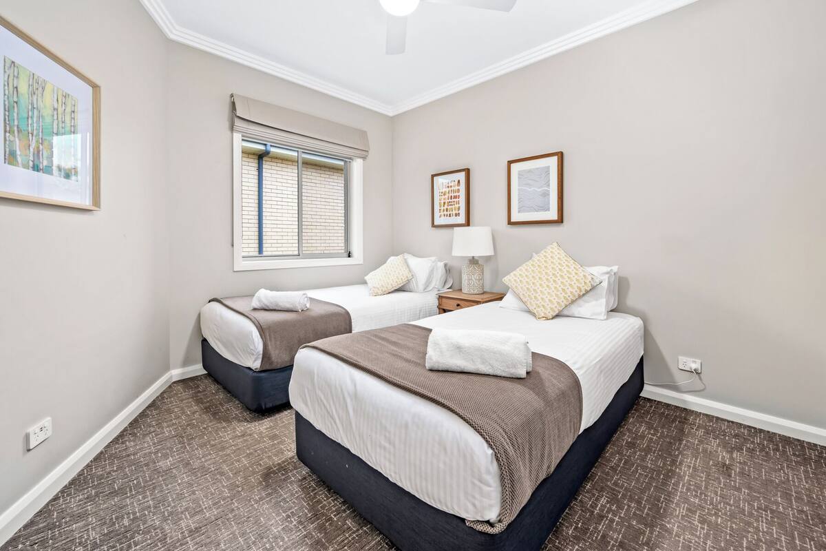 A hotel room with two beds, a window with blinds, a nightstand with a lamp, and artwork on the walls.