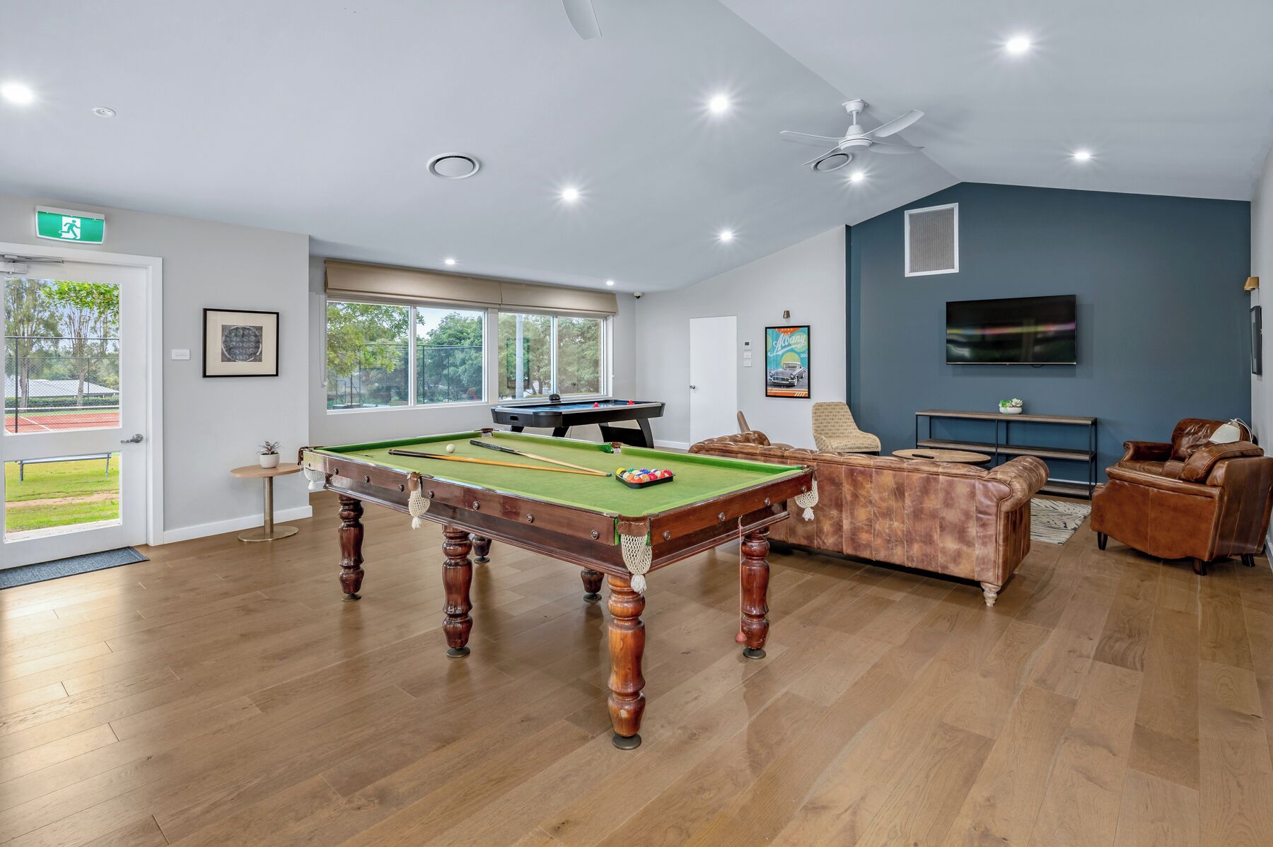 A pool table in a room with wooden flooring, a ceiling fan, and a television mounted on the wall.