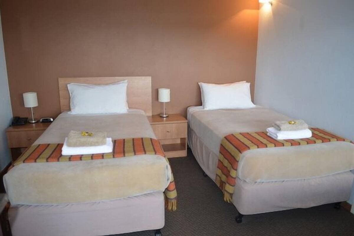 A hotel room with two beds, each with a pillow, a bedside table with a lamp, and a towel on the bed.
