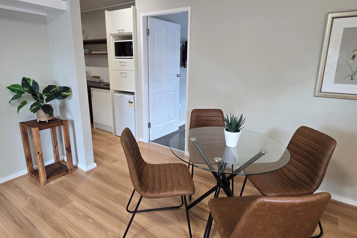 A modern dining area with a glass table and four brown chairs, a wooden side table with a plant, and a kitchen in the background.