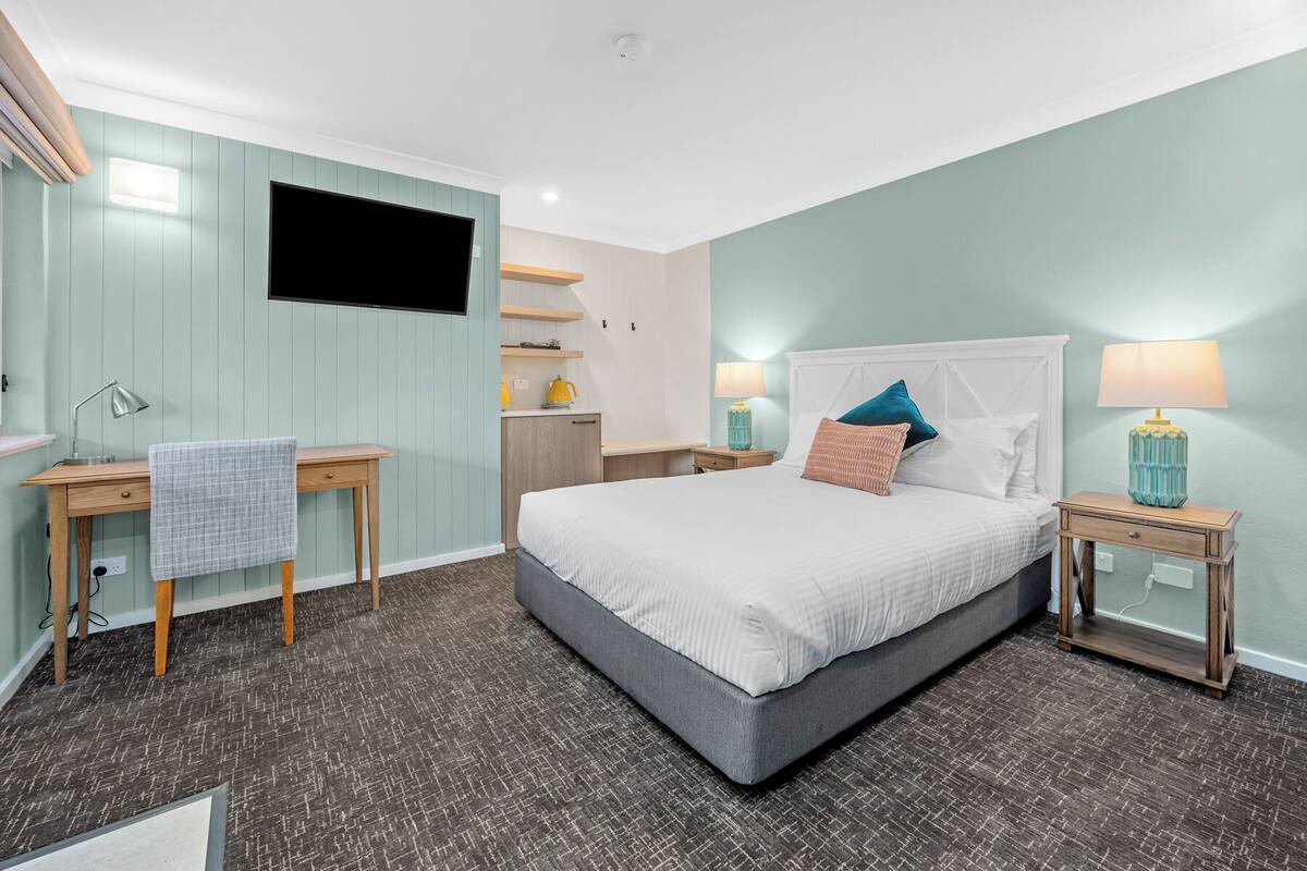 A bedroom with a bed, bedside tables, a desk, a chair, a TV, and a kitchenette.