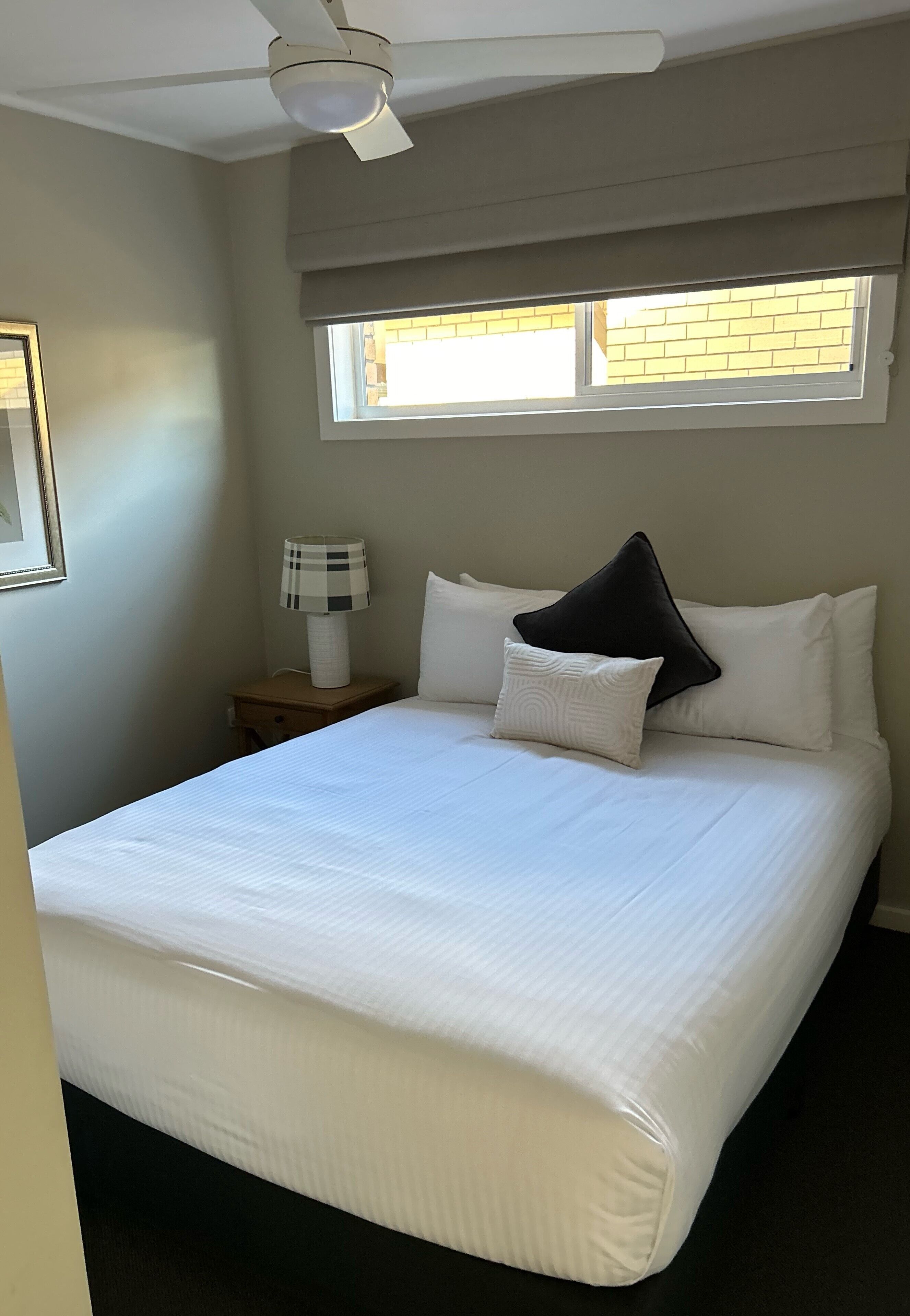 Una cama bien hecha con ropa blanca, una lámpara de noche y una ventana con persianas.