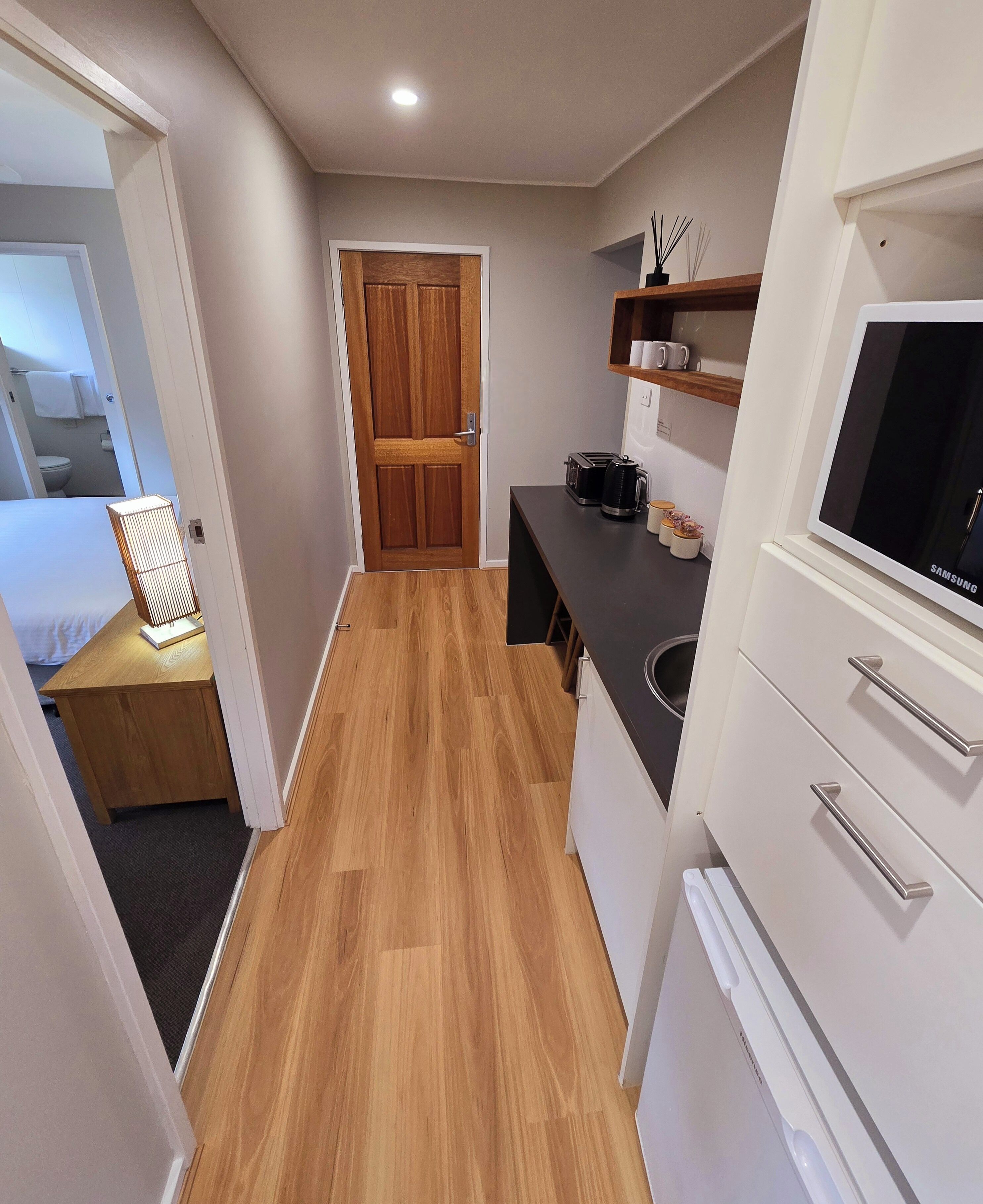 Cocina compacta con piso de madera, horno microondas integrado, televisor Samsung y un pequeño espacio de descanso con mesa y silla.
