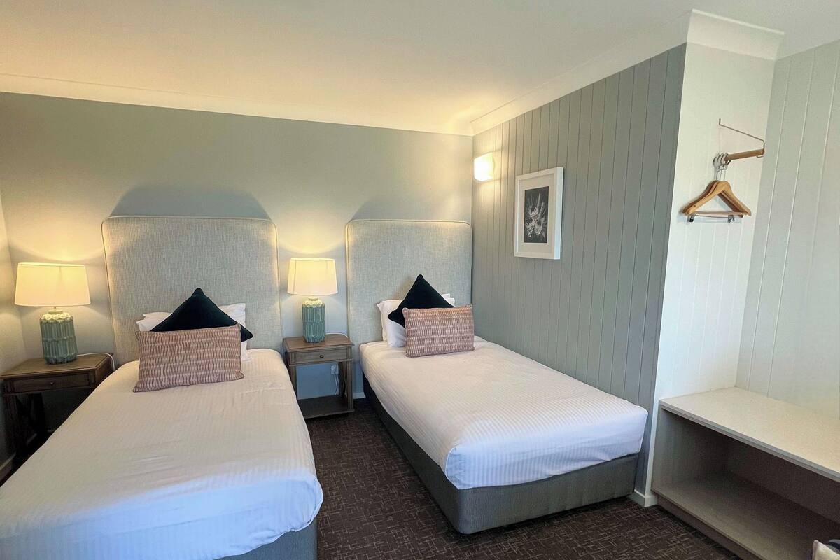 A hotel room with two single beds, nightstands with lamps, a small table, a chair, and a wall-mounted shelf.