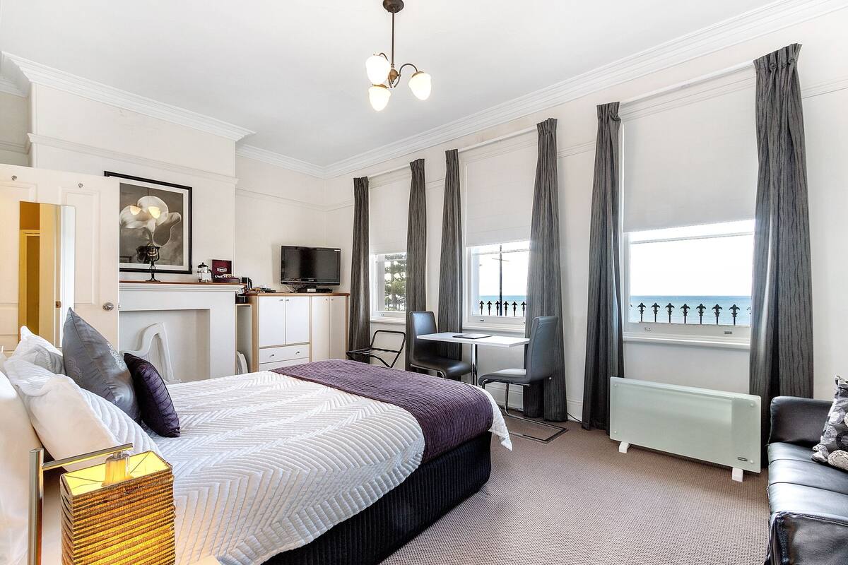 A hotel room with a bed, a desk, a chair, a radiator, and a view of the sea.