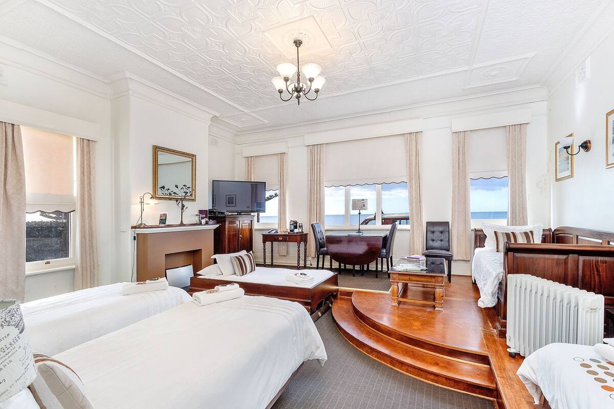 A hotel room with two beds, a desk, a television, and a view of the sea.