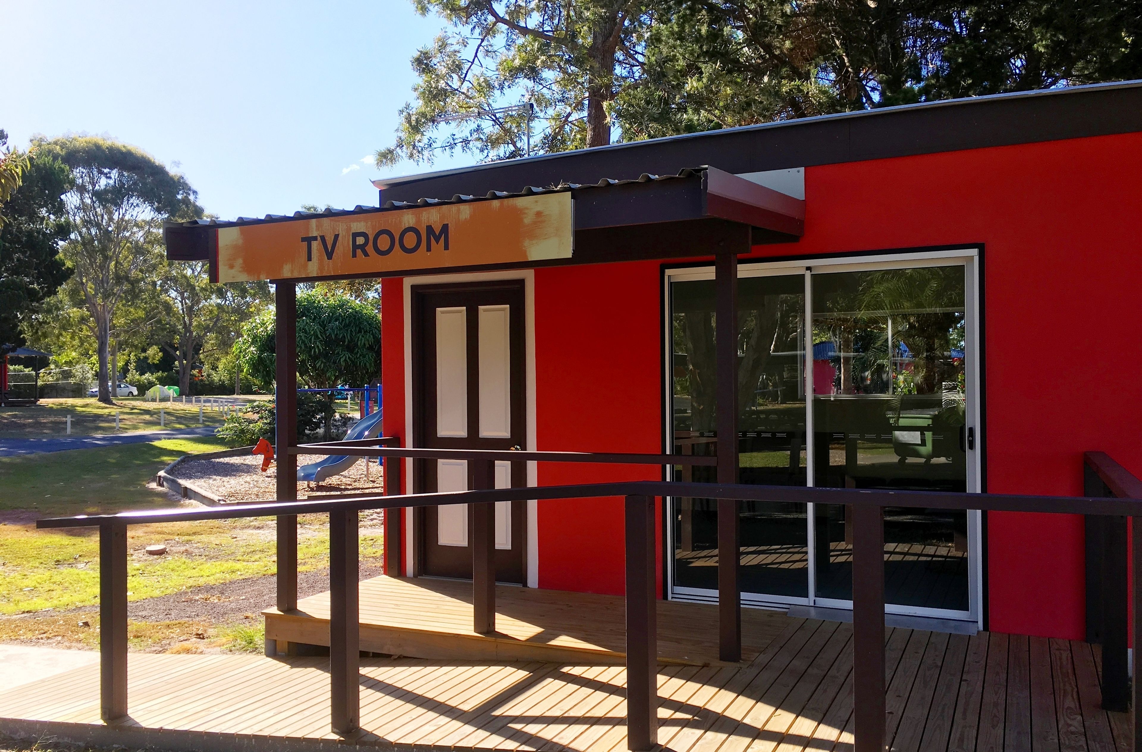 Un bâtiment rouge avec une enseigne indiquant « SALLE DE TÉLÉVISION » et une terrasse en bois menant à l’entrée.