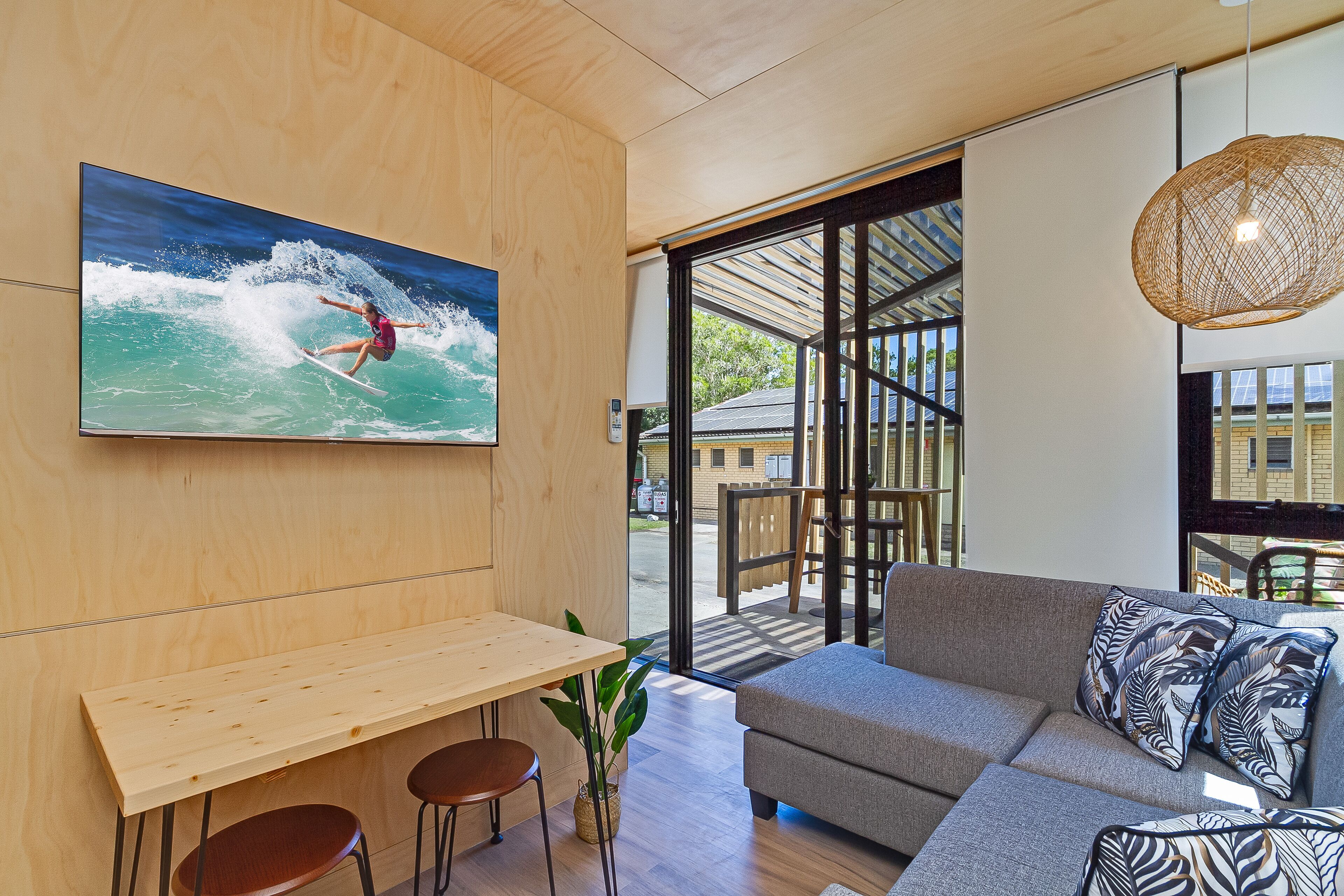 Un salon moderne avec une table en bois, deux tabourets de bar, un canapé gris agr émenté de coussins à motifs et un grand tableau représentant un surfeur sur une vague.