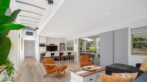 Una sala de estar moderna con un sofá, sillas y una mesita de centro. Se observa una cocina con armarios blancos y un comedor con mesa y sillas.