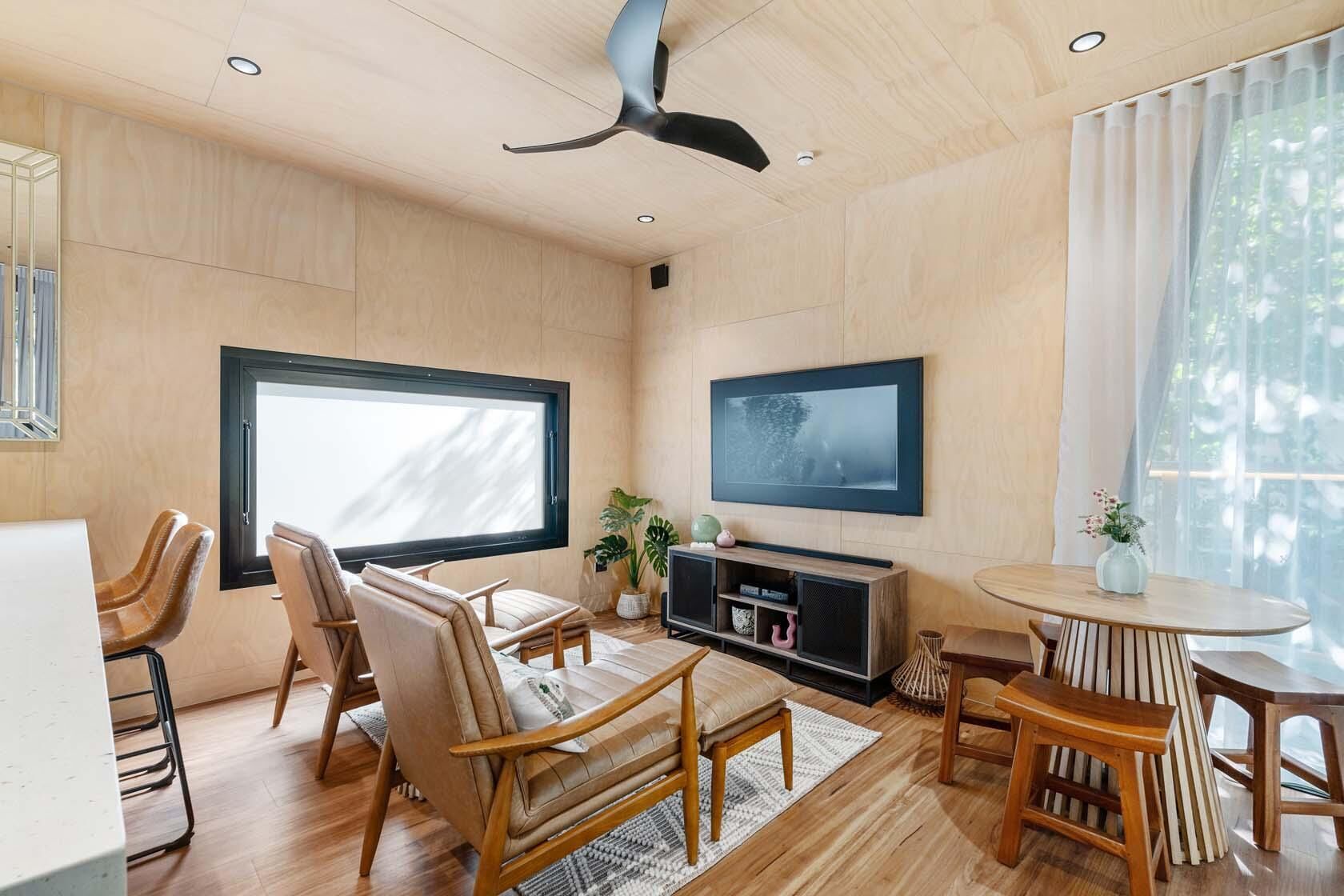 A modern living room with a large window, a flat-screen TV, wooden furniture, and a ceiling fan.