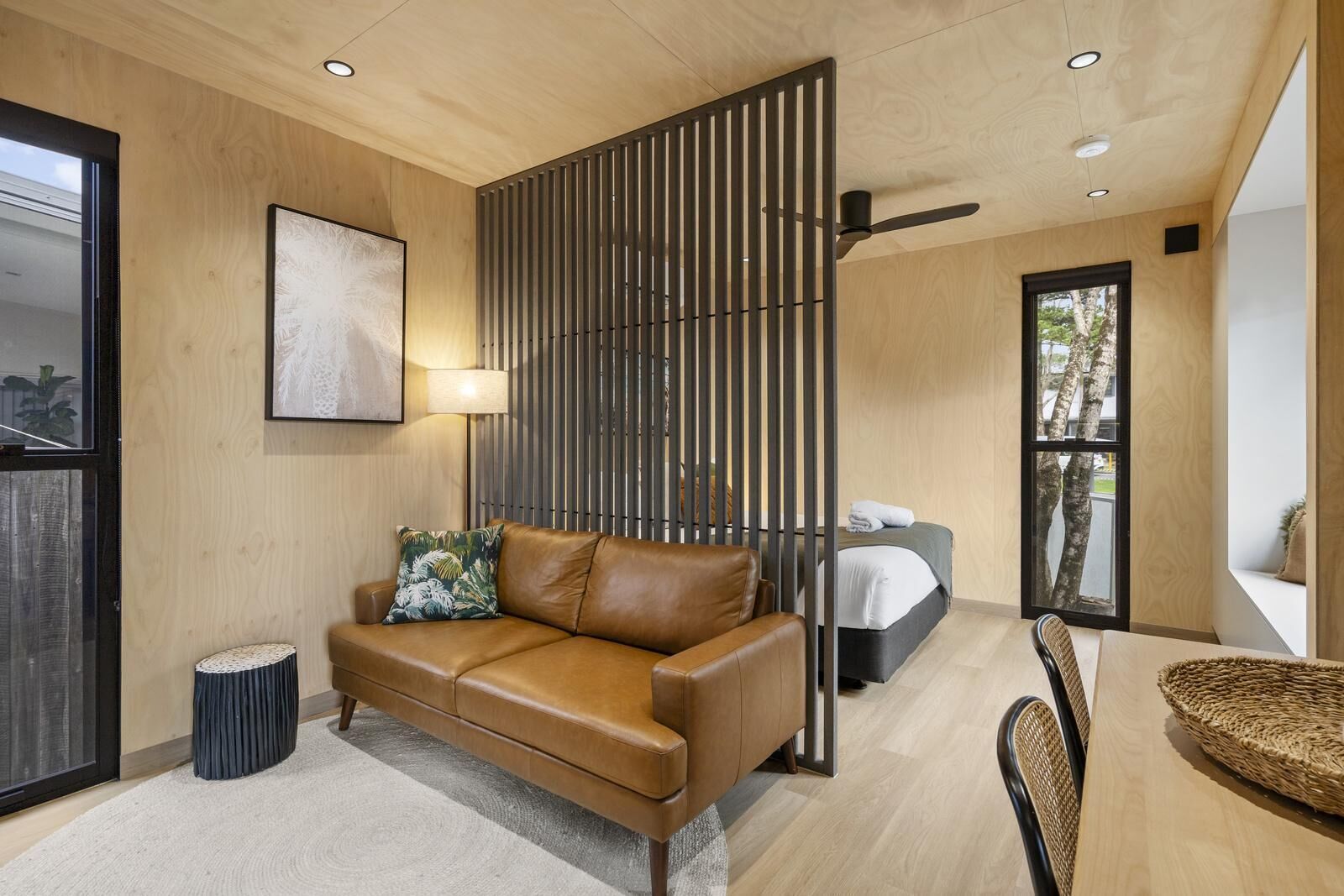 A modern living room with a brown leather sofa, a side table, a lamp, a bed with a white headboard, and a large window.